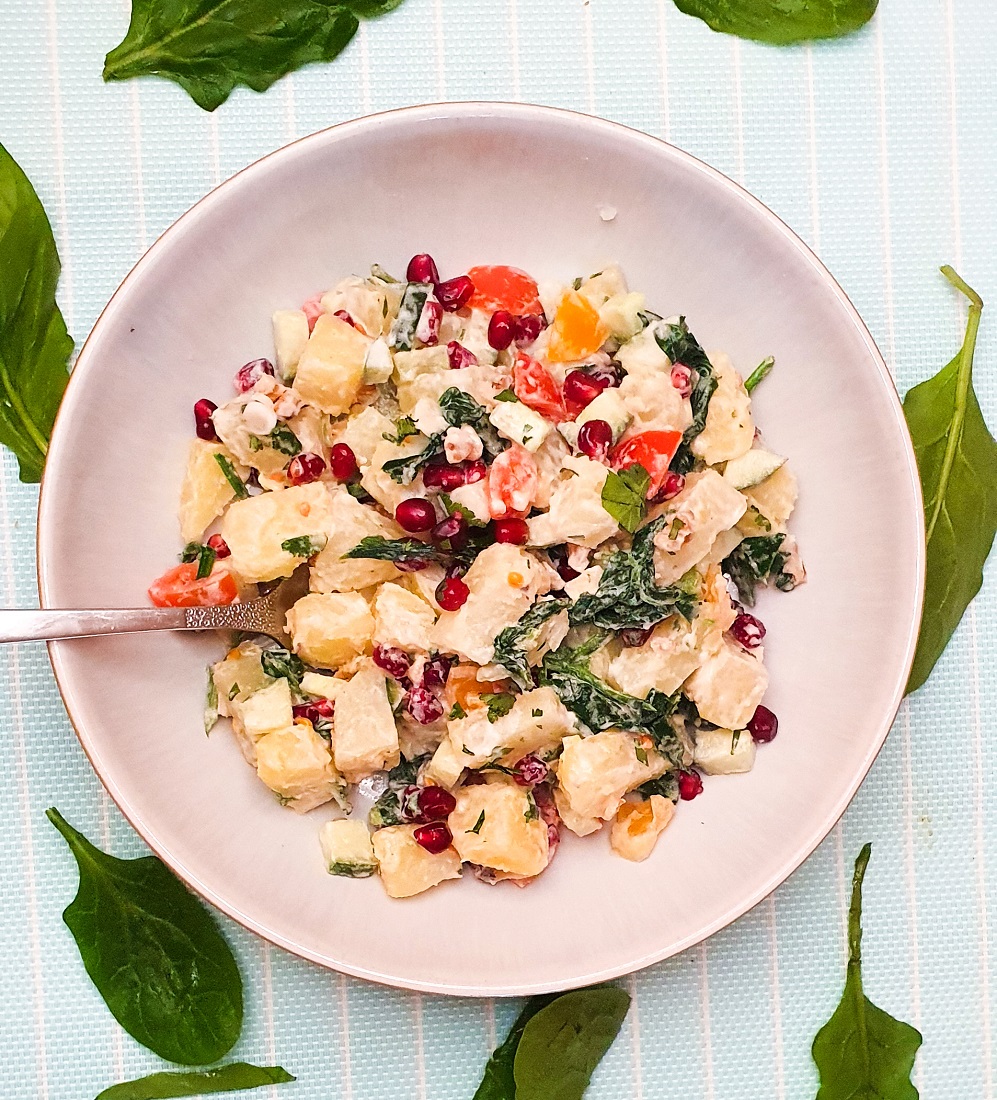 Pomegranate potato salad in a white bowl on a striped blue background with spinach leaves