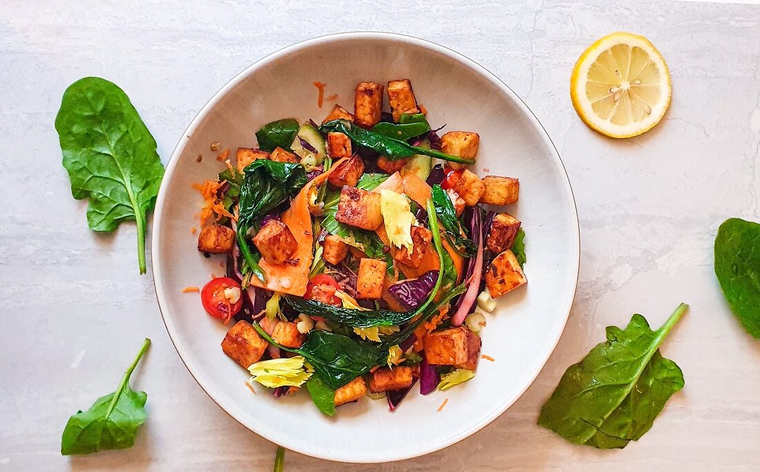 marinaded crispy todu on a bed of veggies in a bowl on a white background with green leaves